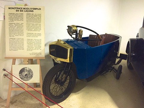 A narrow blue single-track car with a brass radiator, spoked wheels and an outrigger wheel next to a text board on an easel. Opens a larger view