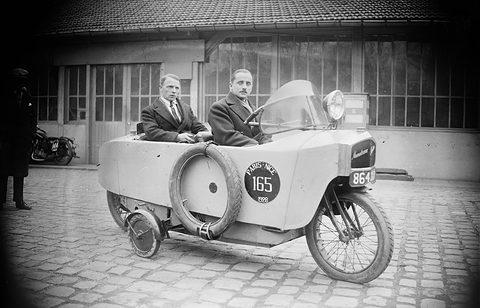 Two men in overcoats sit one behind the other in a narrow car with a spare tyre and a small side wheel.