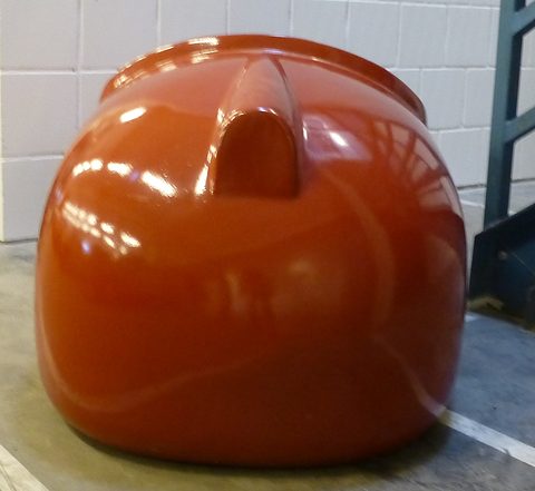 Glossy orange moulded shell with a raised nose hump, standing on a concrete floor by a tiled wall. Opens a larger view