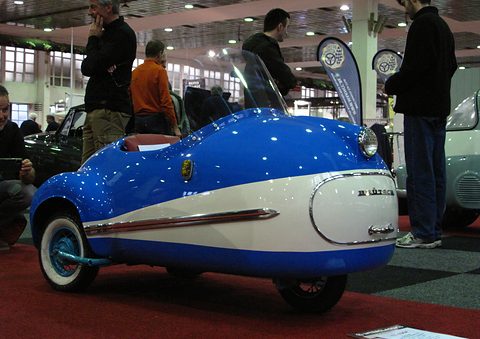 Bright blue single-seater with a white flank and nose panel on a red carpet, visitors standing behind. Opens a larger view