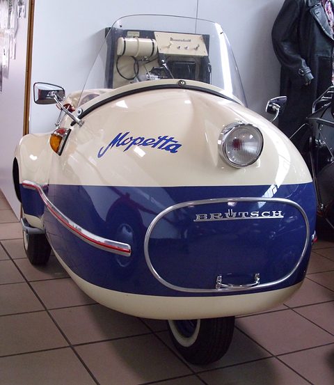 Cream single-seater with a blue lower body, Mopetta script, BRÜTSCH nose panel and chrome mirror on a tiled floor. Opens a larger view