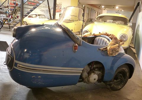 Blue single-seater with its small engine exposed behind the front wheel, cream classic cars behind. Opens a larger view