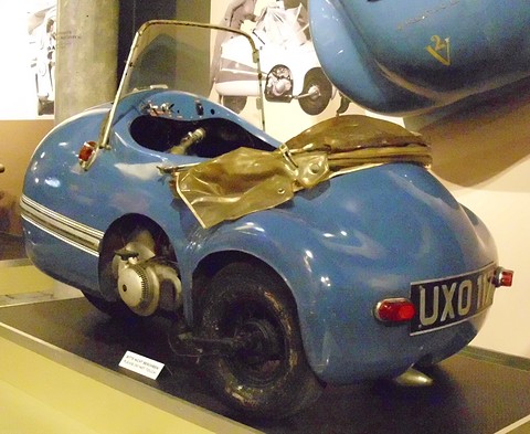 Small blue single-seat three-wheeler from behind, folded hood on its deck and its engine visible beside the rear wheel. Opens a larger view