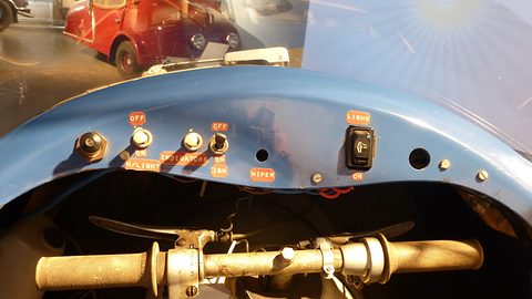 Blue dashboard edge with toggle switches labelled lights, indicators and ignition above a handlebar with grips. Opens a larger view