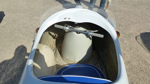 Looking into a white single-seater from above: handlebar on a cream tank and a blue seat cushion, on gravel. Opens a larger view