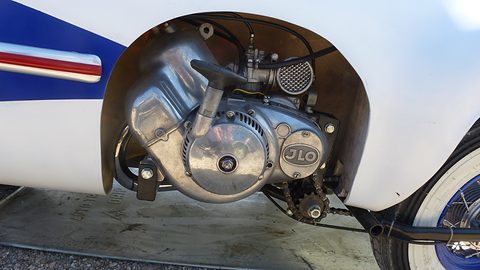 Polished aluminium engine with a JLO cover and mesh air filter in a wheel opening of white bodywork. Opens a larger view