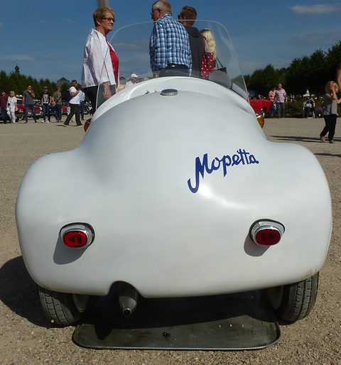 Rounded white tail with two small rear lamps and blue Mopetta script, visitors strolling behind. Opens a larger view