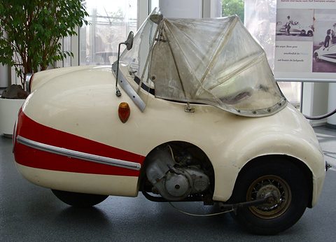 Cream and red single-seat three-wheeler with a clear plastic hood, its engine exposed, beside a potted tree.