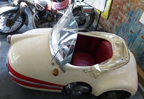 Cream single-seater with a red leather seat, folded clear hood and a spoked front wheel visible beyond. Opens a larger view