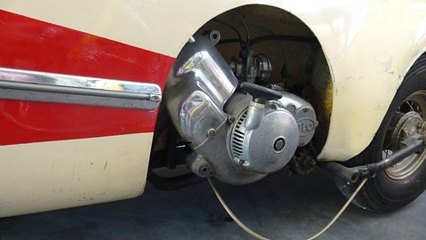 Aluminium single-cylinder engine with cooling grille and chain drive in a wheel opening of cream and red bodywork. Opens a larger view