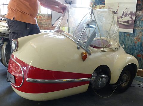 Cream and red single-seater, a man lifting its clear plastic hood, period photographs on the brick wall behind. Opens a larger view
