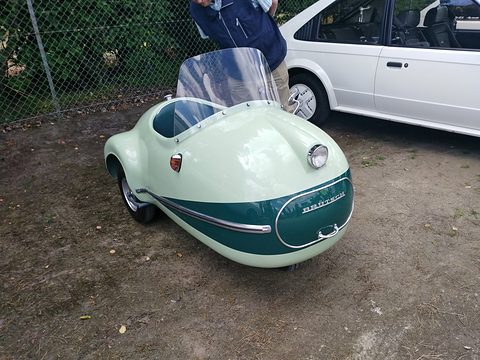 Pale green and dark green single-seat replica with a BRÜTSCH nose badge on bare earth by a fence. Opens a larger view