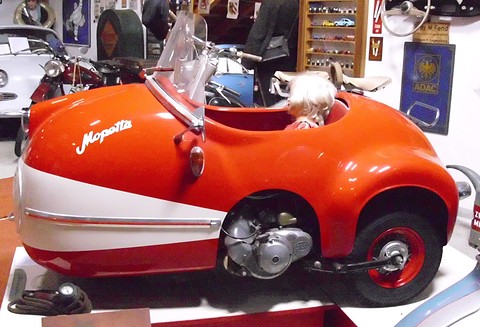 Orange and white single-seat replica with a doll in the seat, enamel signs and model cars behind. Opens a larger view