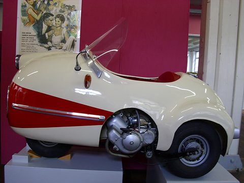 Cream single-seater with red flank and exposed engine, a 1950s film poster on the red wall behind.