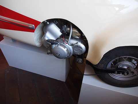 Small silver engine with a pull-start housing beside a spoked wheel, under cream and red bodywork. Opens a larger view