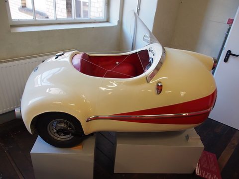 Cream single-seat three-wheeler with a red lower body on a white plinth beneath a window. Opens a larger view