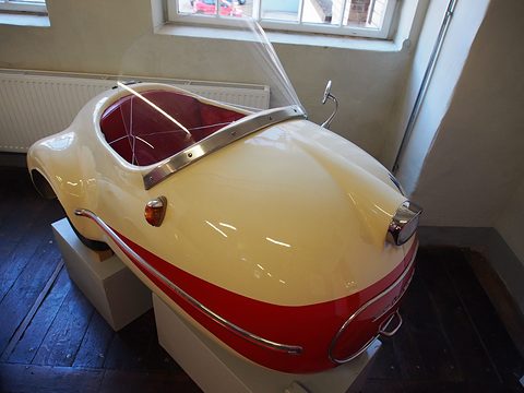 Cream and red single-seater seen from the rear on a plinth on a wooden floor, its windscreen and chrome strip. Opens a larger view