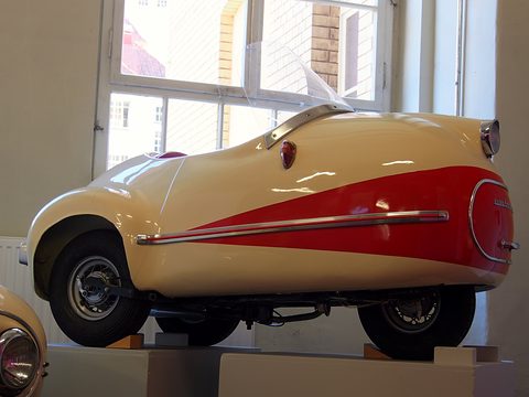 Side view of a cream single-seat three-wheeler with a red sweep along its flank, on a plinth by a window. Opens a larger view