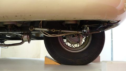 Low view under a cream body: a spoked wheel with drum brake, a coil spring and cables on the frame. Opens a larger view