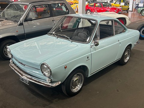 Pale blue small coupé with round headlamps and chrome bumpers on a museum floor, other small cars behind.