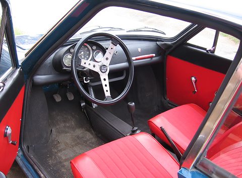 View through the open door: a drilled three-spoke steering wheel, round dials, a floor gear lever and red vinyl seats. Opens a larger view