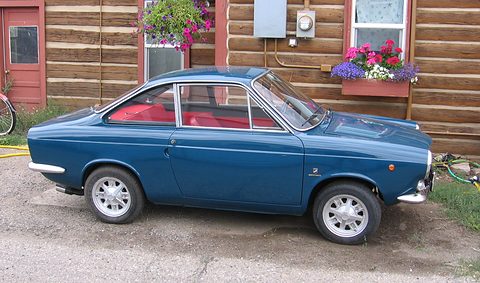 Side view of a dark blue small coupé with red seats and alloy wheels, parked by a log cabin with flower boxes.