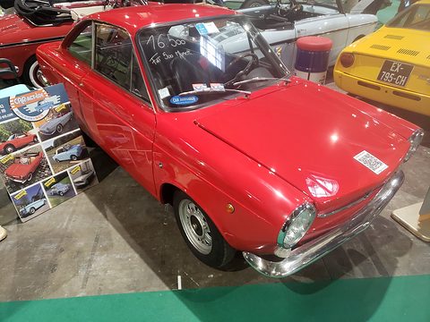 Small red fastback coupé with a sloping bonnet and chrome bumpers on a green show carpet, photo boards beside it. Opens a larger view