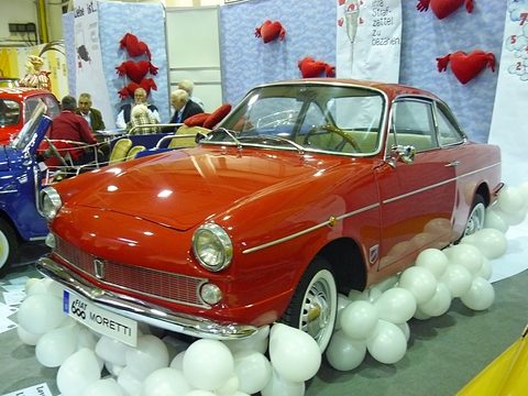 Red two-door coupé with a chrome side strip and a FIAT 600 MORETTI plate, on a stand ringed with white balloons. Opens a larger view