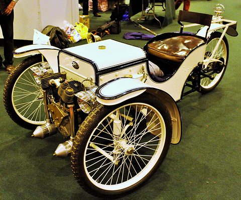 A white single-seat three-wheeler with a box bonnet, brass-trimmed V-twin, buttoned leather seat and wire wheels on green carpet. Opens a larger view