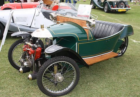 A dark green three-wheeler with wooden side panels, a red-finned V-twin at the nose and a black buttoned seat on grass. Opens a larger view