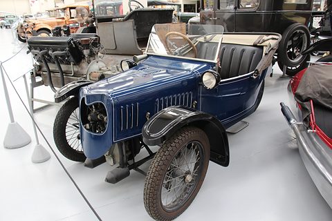 A dark blue three-wheeler with a louvred bonnet, black wings and spoked wheels among other veteran cars in a hall. Opens a larger view