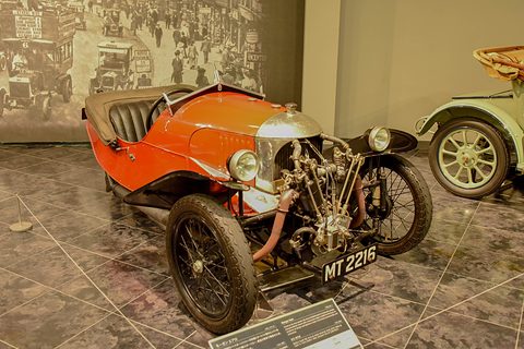 An orange-red three-wheeler with a polished cowl and exposed V-twin before a large period street photograph. Opens a larger view