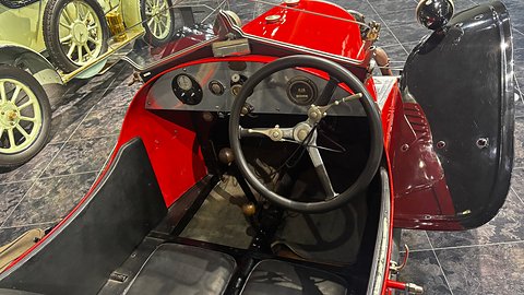 Looking into a red three-wheeler: black steering wheel, small dials on a dark dash and black seat cushions. Opens a larger view