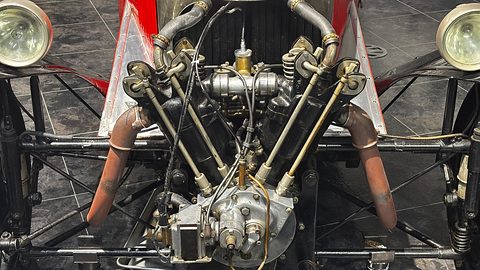 Close front view of an air-cooled V-twin with pushrods and exhaust pipes between two headlamps on a tubular frame. Opens a larger view