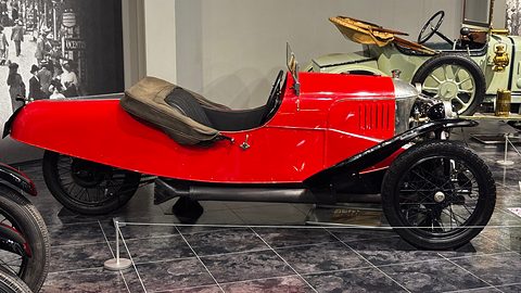 Side view of a red three-wheeler with a pointed tail, louvred bonnet and black wings, a period street photograph behind. Opens a larger view