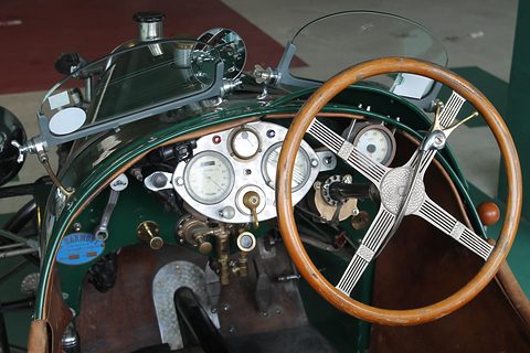 A wooden-rimmed steering wheel with engine-turned spokes before an aluminium panel of round gauges and brass taps. Opens a larger view