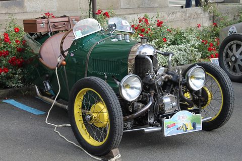 A green three-wheeler with an exposed V-twin and twin headlamps, yellow wire wheels and a rally plate, before rose bushes. Opens a larger view