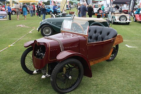 A maroon three-wheeler with a louvred bonnet, buttoned black seat and wooden rear deck on a grass show field. Opens a larger view