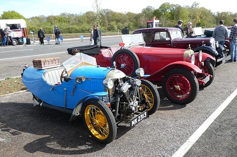 A bright blue three-wheeler with yellow wheels and a hamper on its tail, a red saloon parked beside it. Opens a larger view