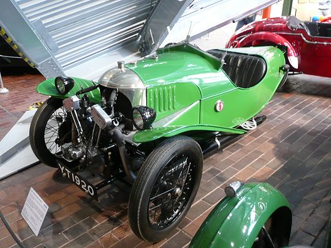 A green three-wheeler with a louvred bonnet and black seats on a brick floor, a placard beside it. Opens a larger view
