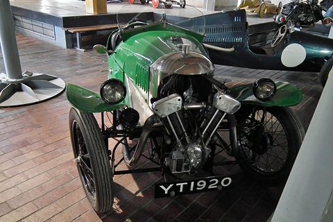 A green three-wheeler with an exposed V-twin and two headlamps on a brick floor, viewed from the front and above. Opens a larger view
