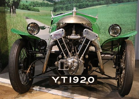 Front view of a green three-wheeler with an exposed V-twin, two headlamps and a number plate, a painted landscape behind. Opens a larger view