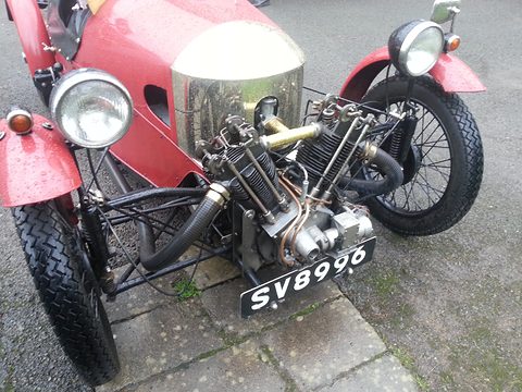 Close front view of a red three-wheeler: two cylinders of a V-twin, a headlamp, coil-sprung pillars and a number plate. Opens a larger view