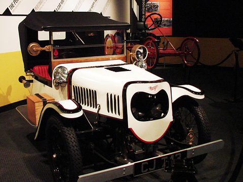 A white three-wheeler with a red-lined bonnet cowl, twin headlamps, a black hood and a long bumper bar. Opens a larger view