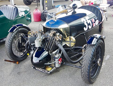 Looking down on a black three-wheeler with an exposed V-twin, exhaust pipes and two headlamps, on tarmac. Opens a larger view