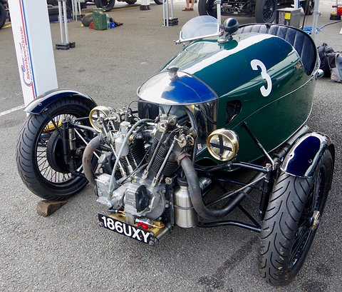 A dark green three-wheeler with a polished cowl, exposed V-twin and racing number 3 on tarmac, gazebos behind. Opens a larger view