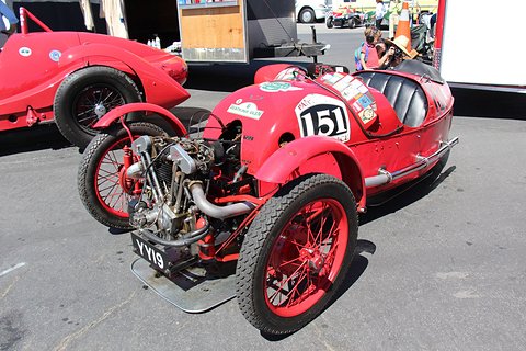 A red three-wheeler with red wire wheels, exposed V-twin and racing number 151, a red racing car behind. Opens a larger view