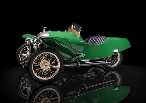 A green three-wheeler with cream wire wheels and a polished cowl on a reflective black floor. Opens a larger view