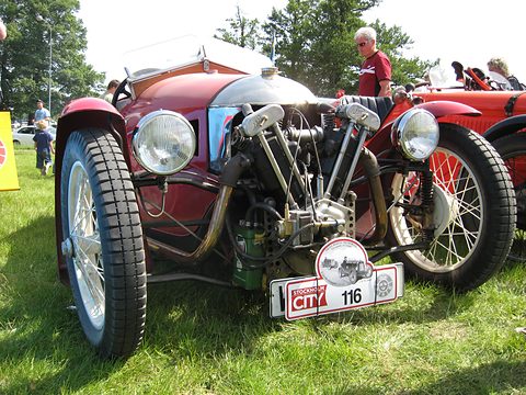 A red three-wheeler with a polished cowl, an exposed V-twin and two headlamps on grass, a rally plate below. Opens a larger view
