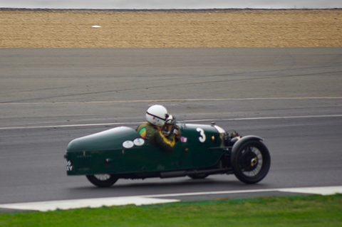 A green streamlined three-wheeler with a helmeted driver on a grey track, a ploughed field behind. Opens a larger view